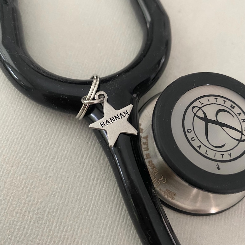May include: A black stethoscope with a silver star charm attached. The charm has the name "HANNAH" engraved on it. The stethoscope has a Littmann brand name on the head.