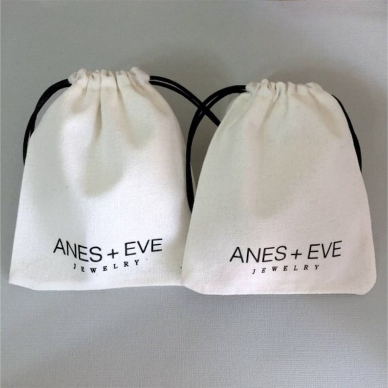 以下が含まれることがあります： 黒い紐の付いた白い綿の巾着袋が2つ。袋には「ANES + EVE JEWELRY」の文字が印刷されています。
