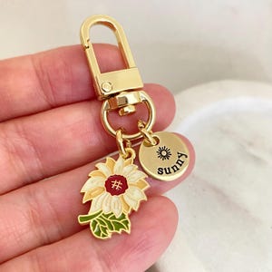 Pode incluir: Chaveiro dourado com um charme de girassol e um charme redondo com a palavra "sunny" e um símbolo de sol.