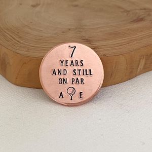May include: A round, copper-colored golf ball marker with the text "7 YEARS AND STILL ON PAR" and a golf ball and tee graphic. The marker also has the letters "A" and "E" on either side of the golf graphic.