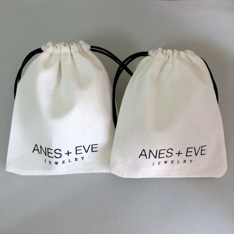 Op de afbeelding: Twee witte trekkoordtassen met zwarte trekkoorden. Beide tassen hebben de tekst "ANES + EVE JEWELRY" in zwart gedrukt.