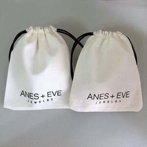 May include: Two white cotton drawstring bags with black drawstrings. The bags have the text "ANES + EVE JEWELRY" printed on the front.