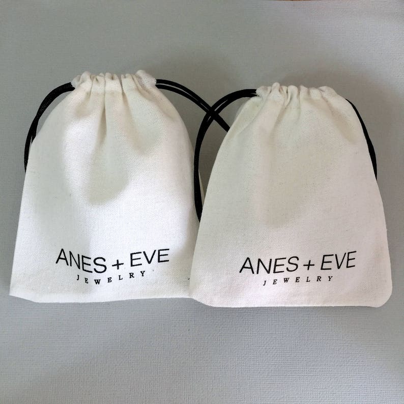 May include: Two white cotton drawstring bags with black drawstrings. The bags have the text "ANES + EVE JEWELRY" printed on the front.