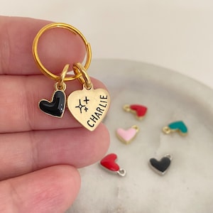 Puede incluir: Llavero dorado con dos colgantes en forma de corazón y un pequeño corazón negro. Un colgante tiene el nombre "CHARLIE" y símbolos de estrellas. Pequeños colgantes de corazón en rojo, rosa y turquesa.