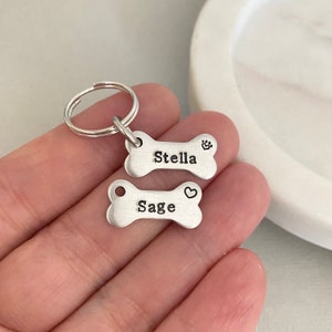 May include: Two silver bone-shaped dog tags with key rings. The top tag says "Stella" with a paw print symbol. The bottom tag says "Sage" with a heart symbol.