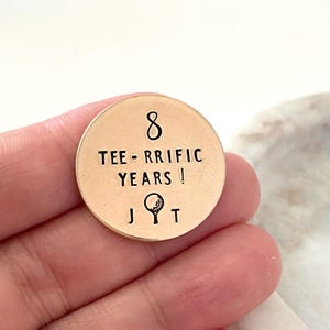 May include: A circular, gold-toned golf ball marker with the inscription "8 TEE-RRIFIC YEARS!" and a golf ball and tee illustration. The marker also features the initials "J T". The marker is held in a hand.