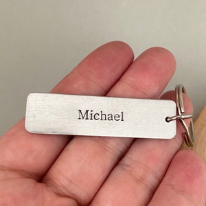 K&ouml;nnte beinhalten: Silberner Schl&uuml;sselanh&auml;nger mit geb&uuml;rsteter Metalloberfl&auml;che und dem eingravierten Namen "Michael".