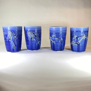 May include: Four blue ceramic tumblers with a textured, ribbed design. Each tumbler features a raised relief of a dolphin. The dolphins are a lighter shade of blue, creating a contrast against the darker blue background.