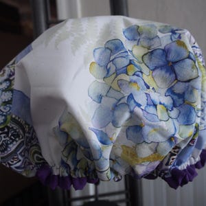 Puede incluir: Un gorro de ducha blanco con un diseño floral de hortensias azules y amarillas. El gorro tiene un ribete fruncido morado en el borde. El diseño también incluye un estampado de cachemira en tonos azules, verdes y morados.