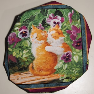 Puede incluir: Dos gatitos naranja y blancos se acurrucan en un jardín con flores moradas y blancas. Los gatitos están sobre una superficie de madera con hojas verdes y flores detrás de ellos.