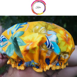Puede incluir: Un gorro de ducha amarillo con un estampado floral tropical con flores de hibisco y hojas de palmera en tonos amarillos, azules y verdes. El gorro tiene un borde con volantes y un cordón. El logo "Kehni" está en un círculo colorido.