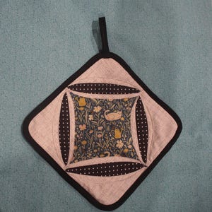 Peut inclure: Un dessous de plat matelassé avec une bordure noire et une boucle de suspension. Le centre présente un motif floral avec un chat et des arrosoirs. Le design comprend des accents de pois noirs et un fond gris clair.