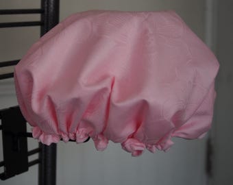 May include: A light pink shower cap with a floral pattern. The cap is gathered at the base with a ruffled edge. The fabric has a subtle sheen. The shower cap is hanging on a black metal rack.