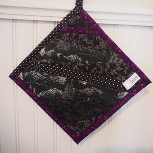 May include: A quilted, diamond-shaped pot holder featuring a black and white mountain scene, leaf patterns, and polka dots. It has a purple border and a hanging loop. The label reads "Québec".