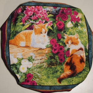 Puede incluir: Una tela colorida con un diseño floral que presenta dos gatos atigrados naranja y blanco. Los gatos están sentados en un jardín con flores rosas y follaje verde.