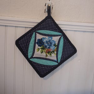 Peut inclure: Un dessous de plat matelassé avec un motif à pois bleu marine et une bordure turquoise. Le centre présente un motif floral avec des fleurs bleues et des feuilles vertes. Le dessous de plat est accroché à un mur blanc.