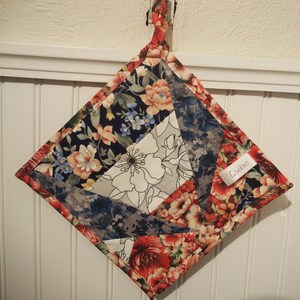 Peut inclure: Un dessous de plat matelassé en forme de losange avec un motif floral rouge, bleu et blanc. Le dessous de plat a une boucle pour l'accrocher et une petite étiquette avec le mot "Québec".