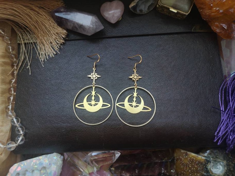 May include: Gold tone earrings featuring a crescent moon and star design. The earrings are drop earrings and have a circular shape.