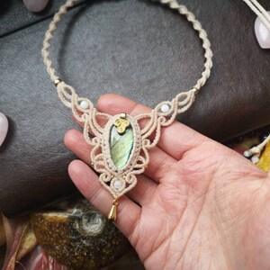 Puede incluir: Un delicado collar de macram&eacute; con un colgante de labradorita verde de forma ovalada. El collar est&aacute; hecho con cord&oacute;n de color crema e incluye peque&ntilde;as cuentas blancas.