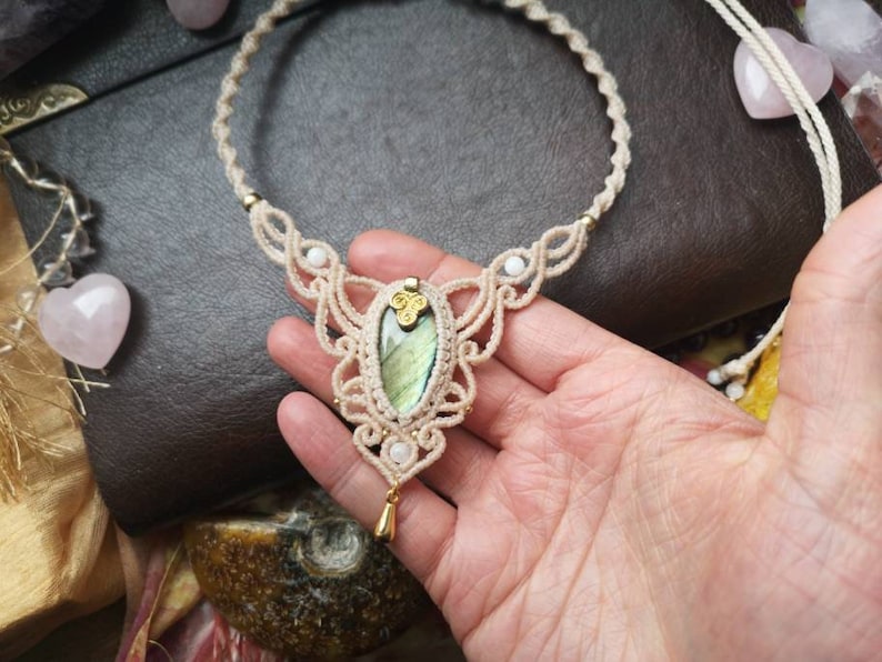 Puede incluir: Un delicado collar de macram&eacute; con un colgante de labradorita verde de forma ovalada. El collar est&aacute; hecho con cord&oacute;n de color crema e incluye peque&ntilde;as cuentas blancas.