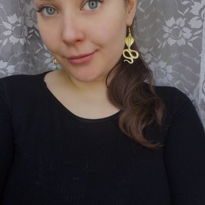 May include: A woman wearing a black shirt with gold snake earrings. The earrings are dangling and feature a detailed snake design.