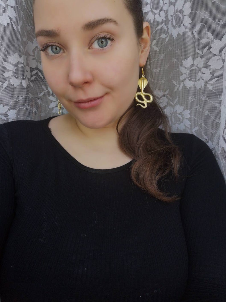 May include: A woman wearing a black shirt with gold snake earrings. The earrings are dangling and feature a detailed snake design.