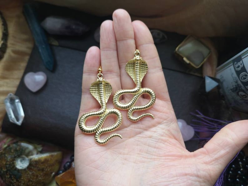 May include: A pair of gold-tone snake earrings. Each earring features a detailed cobra design with a raised, textured surface.