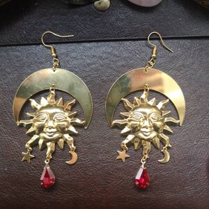 May include: Gold earrings with a sun and crescent moon design. Each earring features a detailed sun face with a red gemstone dangle.
