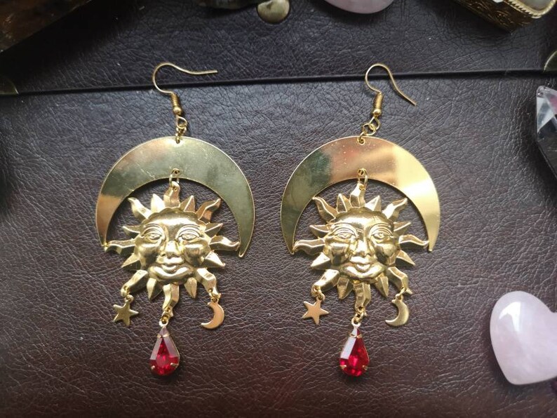 May include: Gold earrings with a sun and crescent moon design. Each earring features a detailed sun face with a red gemstone dangle.