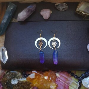 Puede incluir: Un par de pendientes de oro con un dise&ntilde;o de luna creciente y un amuleto de mano de hamsa. Cada pendiente presenta un colgante de cristal morado y azul.