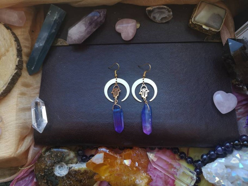 Puede incluir: Un par de pendientes de oro con un dise&ntilde;o de luna creciente y un amuleto de mano de hamsa. Cada pendiente presenta un colgante de cristal morado y azul.