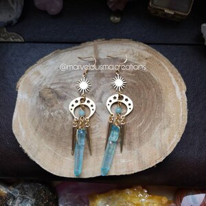 May include: Gold earrings with a crescent moon and star design, featuring a blue crystal pendant. The earrings are on a wooden surface.