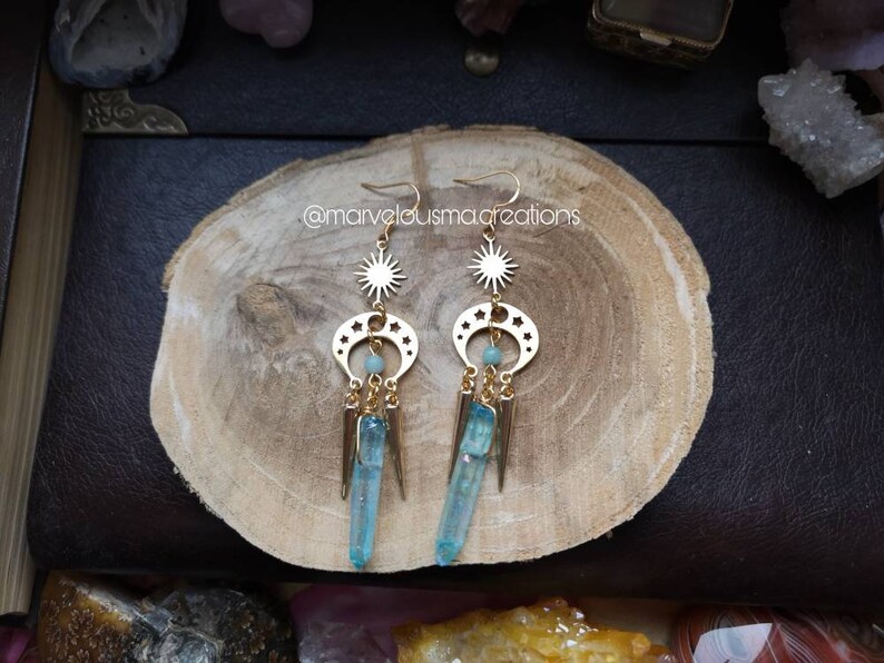 May include: Gold earrings with a crescent moon and star design, featuring a blue crystal pendant. The earrings are on a wooden surface.