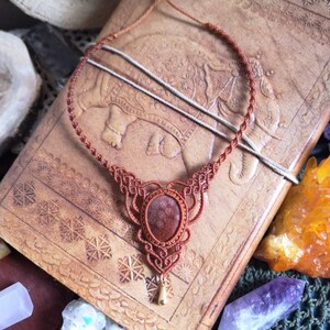 Puede incluir: Un collar de cord&oacute;n de cuero marr&oacute;n con un colgante de piedra roja. El collar est&aacute; anudado en un patr&oacute;n decorativo y tiene un peque&ntilde;o dije de oro en la parte inferior.