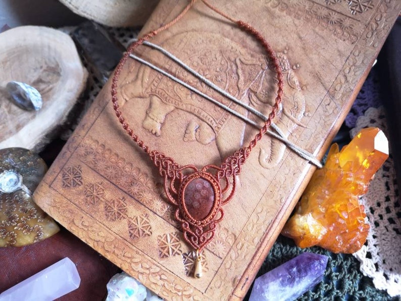 Puede incluir: Un collar de cord&oacute;n de cuero marr&oacute;n con un colgante de piedra roja. El collar est&aacute; anudado en un patr&oacute;n decorativo y tiene un peque&ntilde;o dije de oro en la parte inferior.