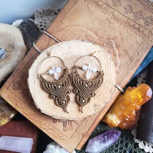 May include: A pair of gold and brown macrame earrings with pink crystal accents. The earrings are displayed on a wooden surface.