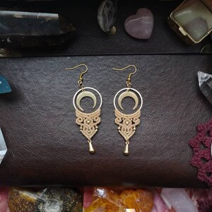 May include: A pair of gold crescent moon earrings with a beige macrame tassel. The earrings are displayed on a brown leather surface with crystals.