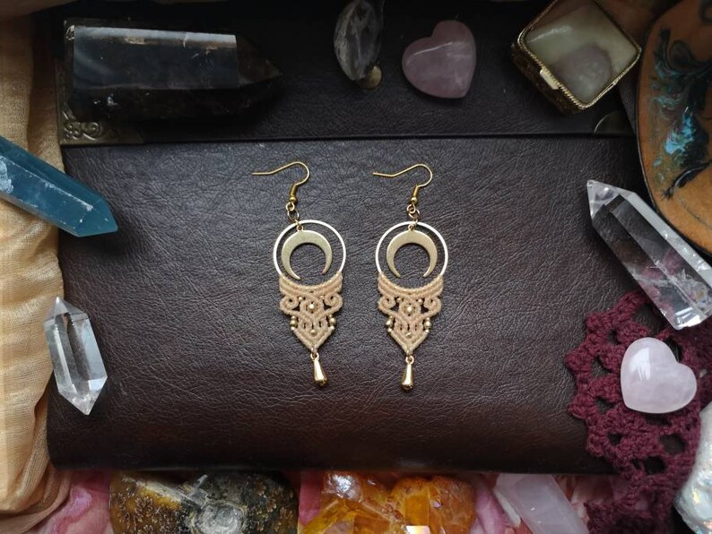 May include: A pair of gold crescent moon earrings with a beige macrame tassel. The earrings are displayed on a brown leather surface with crystals.