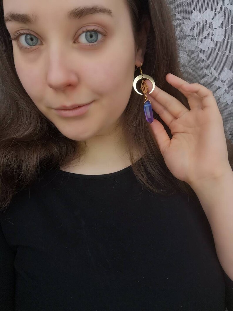 Puede incluir: Pendientes de media luna dorados con un amuleto de mano de Hamsa y un colgante de cristal morado.