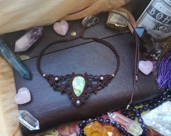 Puede incluir: Un collar de macramé marrón con un colgante de piedra verde y pequeñas cuentas blancas. El collar se muestra en un diario de cuero marrón con cristales y otras joyas.