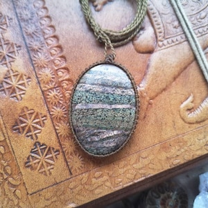 Puede incluir: Un diario de cuero marrón con un colgante ovalado decorativo con una piedra verde y marrón con un brillo metálico.