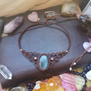 May include: A blue and green gemstone pendant necklace with a macrame design. The necklace is made with brown cord and features a blue and green gemstone pendant. The necklace is displayed on a brown leather surface.