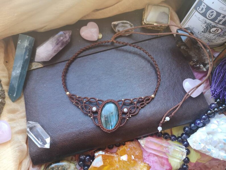 May include: A blue and green gemstone pendant necklace with a macrame design. The necklace is made with brown cord and features a blue and green gemstone pendant. The necklace is displayed on a brown leather surface.