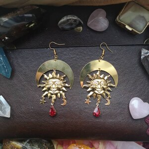 May include: A pair of gold earrings with a crescent moon design and a sun face in the center. Each earring has a red gemstone dangling from the bottom.