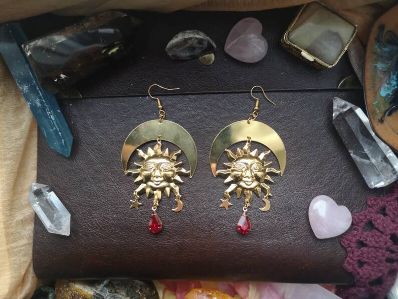 May include: A pair of gold earrings with a crescent moon design and a sun face in the center. Each earring has a red gemstone dangling from the bottom.