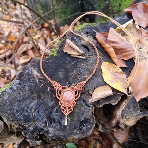 Puede incluir: Un collar de macram&eacute; marr&oacute;n con un colgante de piedra rojo-naranja. El collar se muestra sobre un toc&oacute;n de &aacute;rbol con hojas de oto&ntilde;o.