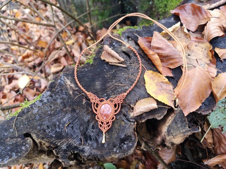 Puede incluir: Un collar de macram&eacute; marr&oacute;n con un colgante de piedra rojo-naranja. El collar se muestra sobre un toc&oacute;n de &aacute;rbol con hojas de oto&ntilde;o.