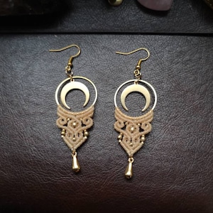May include: A pair of gold crescent moon earrings with a beige macrame design. The earrings have a dangling drop at the bottom.