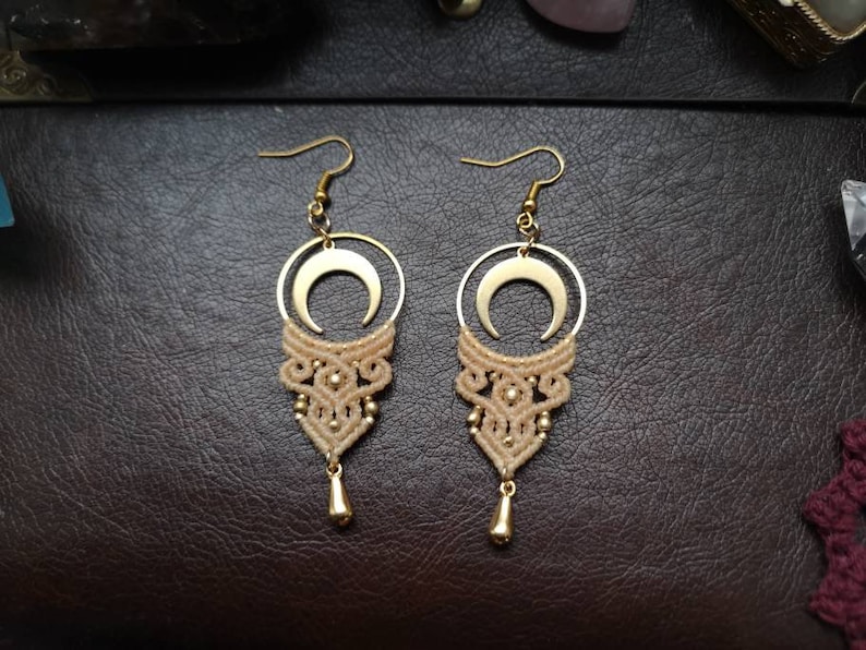 May include: A pair of gold crescent moon earrings with a beige macrame design. The earrings have a dangling drop at the bottom.