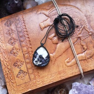 Peut inclure: Un pendentif en forme de larme noir et blanc avec un collier en cordon noir. Le pendentif est fait d'une pierre naturelle avec un motif unique. Le collier est posé sur un journal en cuir marron avec un motif d'éléphant.